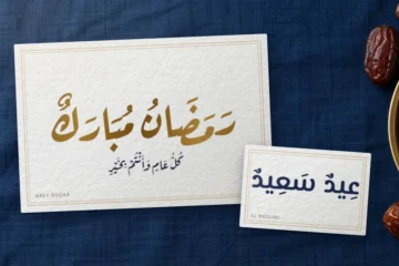 بطاقتا تهنئة على مفرش أزرق داكن: «رمضان مبارك» مذهّبةً بخط رقعة عارف، و«عيد سعيد» بخط المسيري، وبجانبهما طبق تمر نحاسي